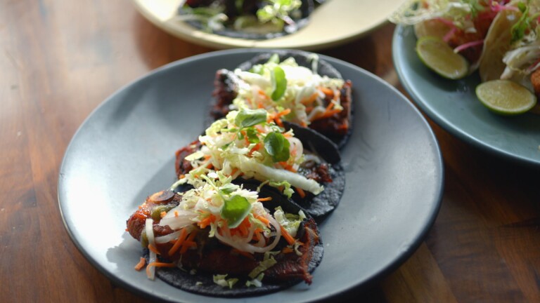 Three tacos are served on a dark gray ceramic plate. The tacos are made of black tortillas and topped with a red Korean short rib and topped with shredded daikon, carrots, napa cabbage and green garnish. The plate sits on a dark wood table and is surrounded by two other plates at the corner of the photo.