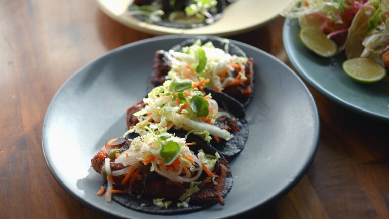 Three tacos are served on a dark gray ceramic plate. The tacos are made of black tortillas and topped with a red Korean short rib and topped with shredded daikon, carrots, napa cabbage and green garnish. The plate sits on a dark wood table and is surrounded by two other plates at the corner of the photo. 