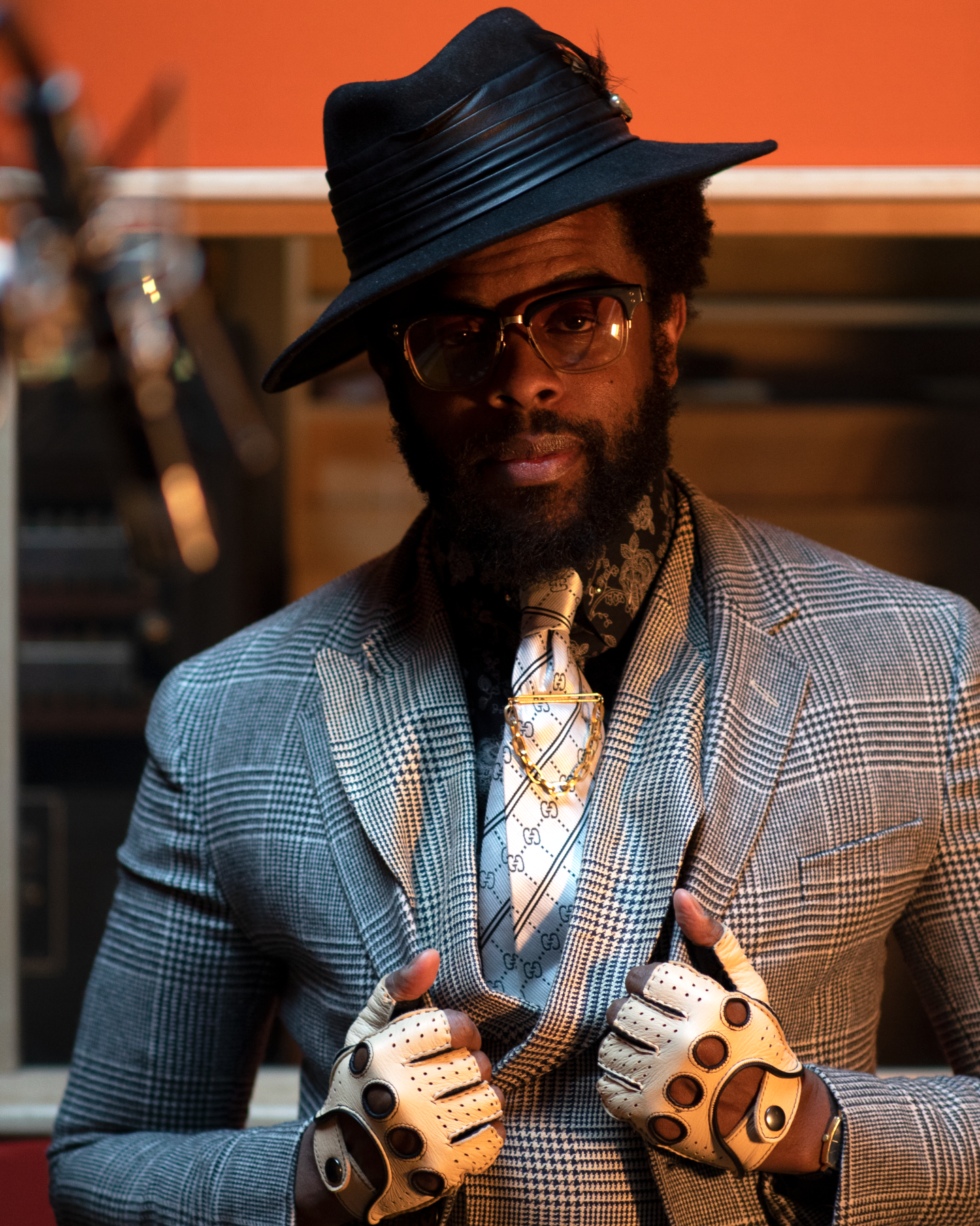 Adrian Younge poses facing the camera. He's wearing a plaid suit jacket and a silk black and white tie. He's also wearing a black brimmed hat tilted slightly to the left. Thick-framed glasses sit on his face and he's wearing white, fingerless gloves.