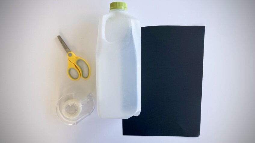 an empty milk jug, tape, scissors and a sheet of black paper on a table