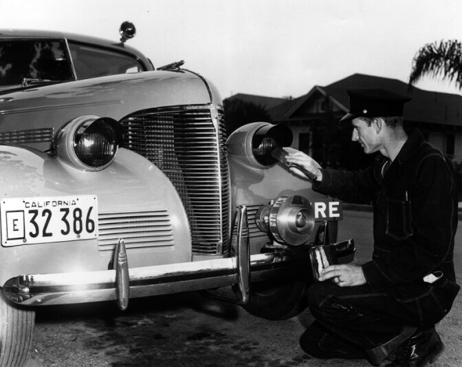 An archival black-and-white photo of a man kneeled with his hand on a vintage car.