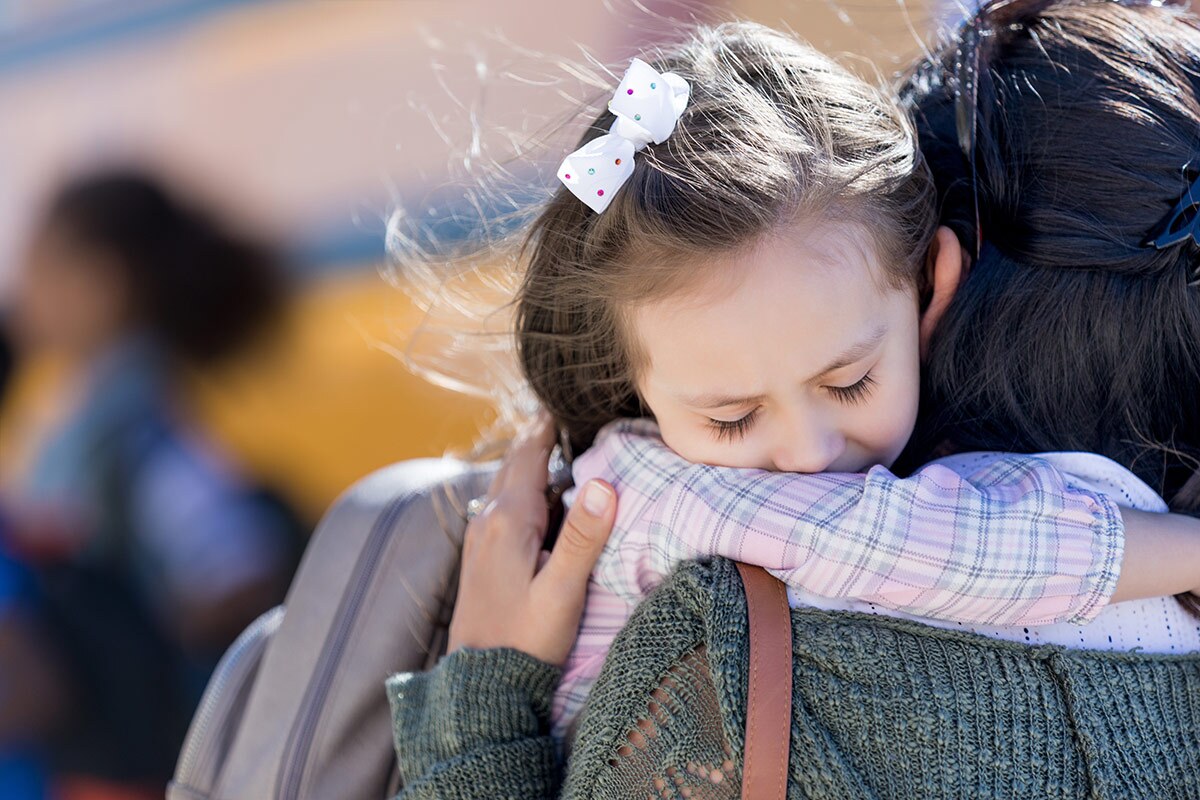 A small child hugs a caregiver goodbye in front of a school bus.