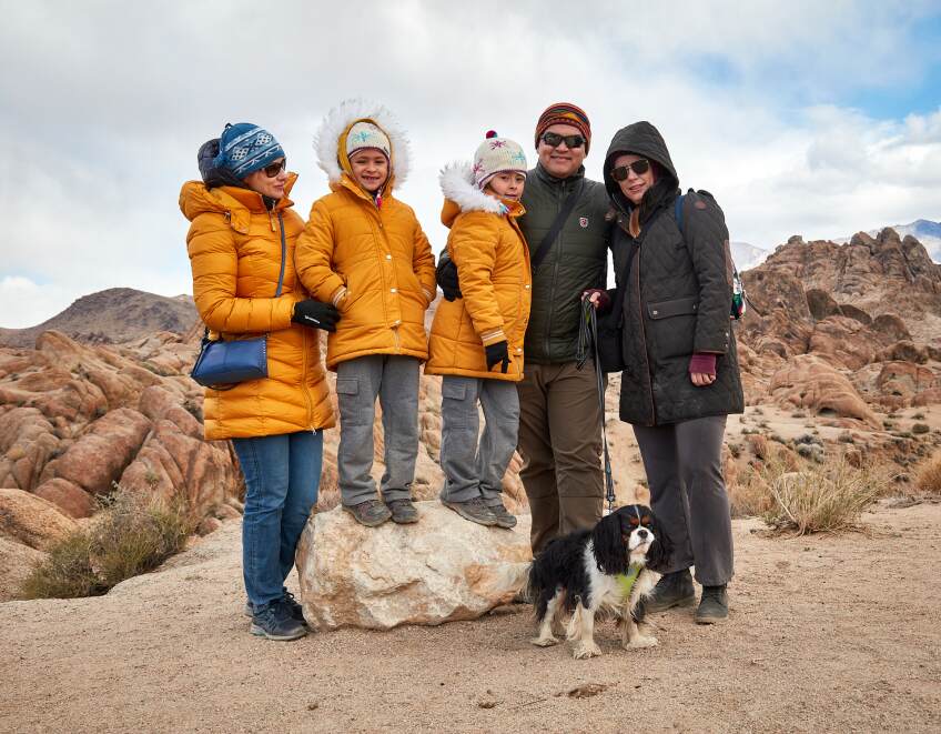 A group family photo taken in the desert. Two children stand on top of a rock, wearing yellow puffer jackets. To their left is a woman wearing a yellow puffer jacket and a blue beanie. To their right are a man and a woman in dark puffer jackets. The man is holding a leash that's attached to a black and white dog standing at their feet.