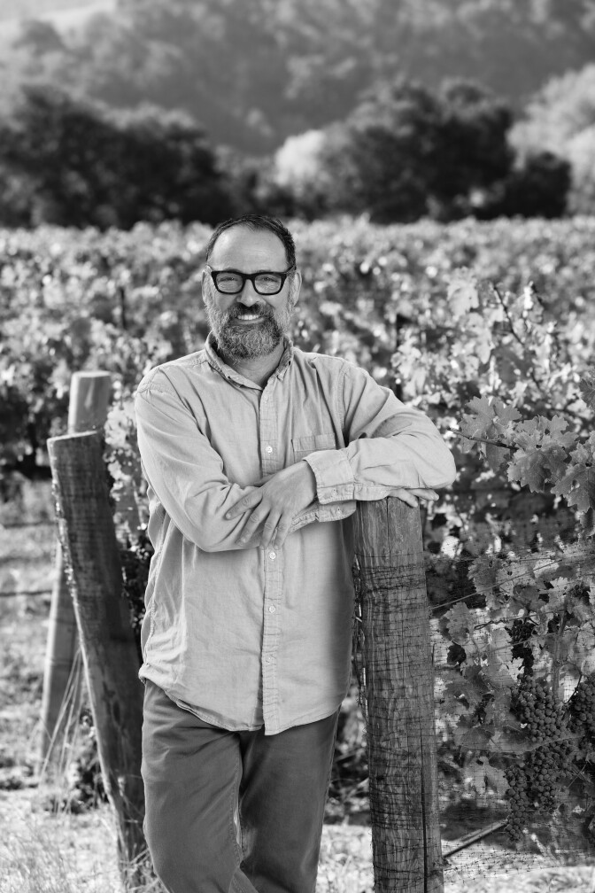 A black and white photo of Larry Schaffer of Tercero Wines standing in a vineyard. He's leaning against a wooden pole and has his arms crossed over his chest. He's smiling and wears a button up shirt and thick-rimmed black glasses.