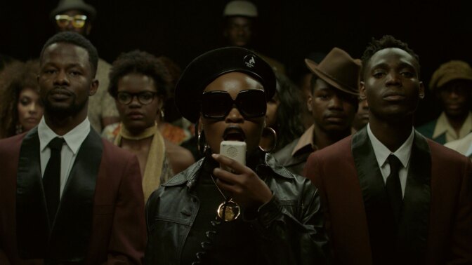 Still from Pongo's "Tamulya": A woman with a beret, leather jacket, sunglasses and a PA microphone, with men in suits on either side and others in the background