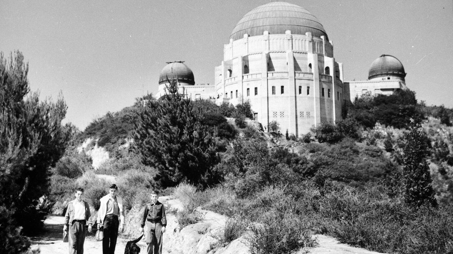A few people trek down a road with the Griffith Observatory in the background.