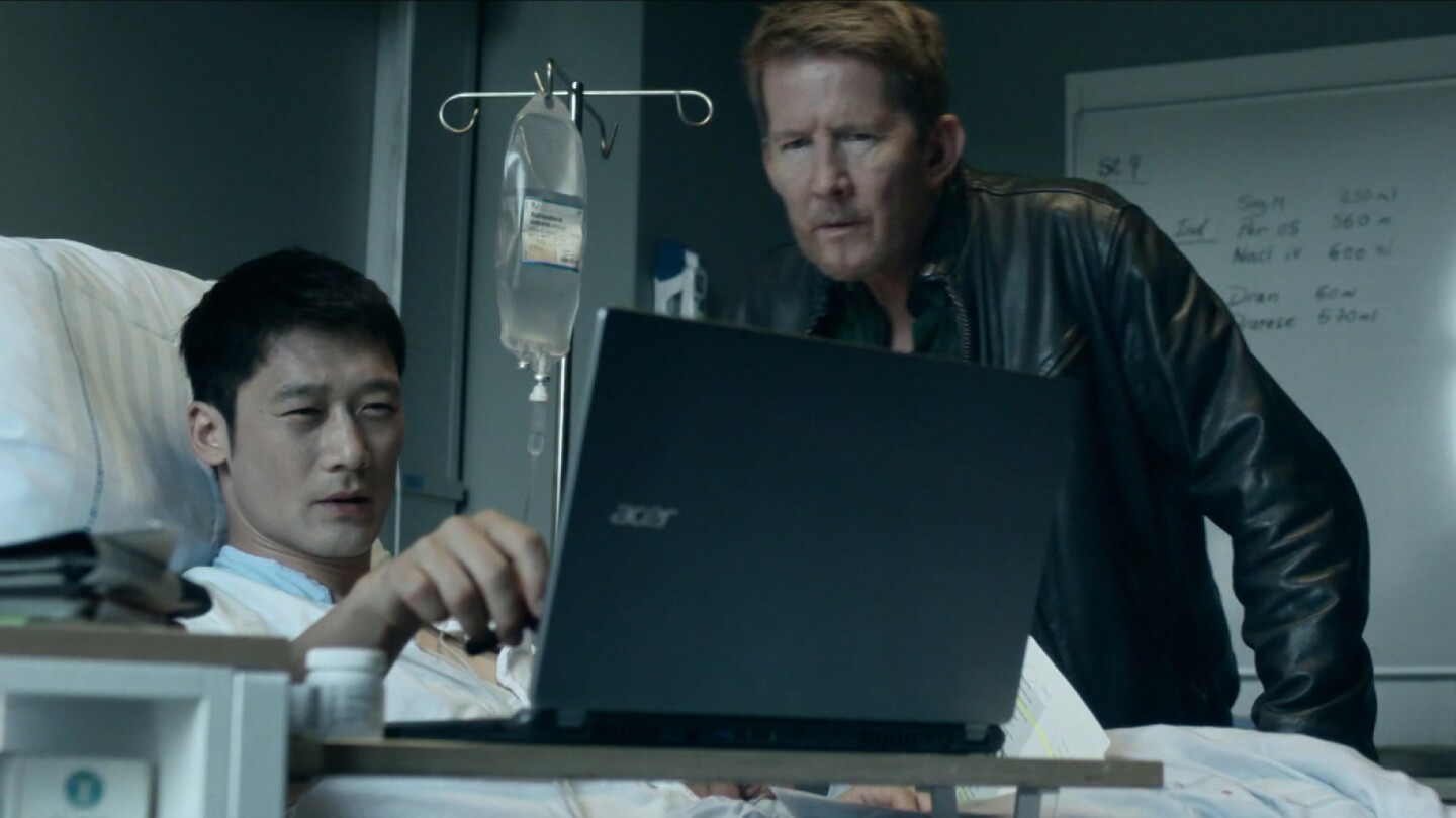 Alf (left) lies in a hospital bed looking at a computer screen with Mads (right) standing next to him. | Still from "Follow the Money," Season 2, Episode 9