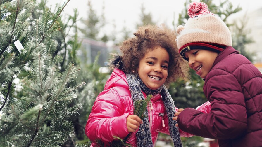 Two smiling children in puffy, colorful coats stand near snowy pine trees. istock
