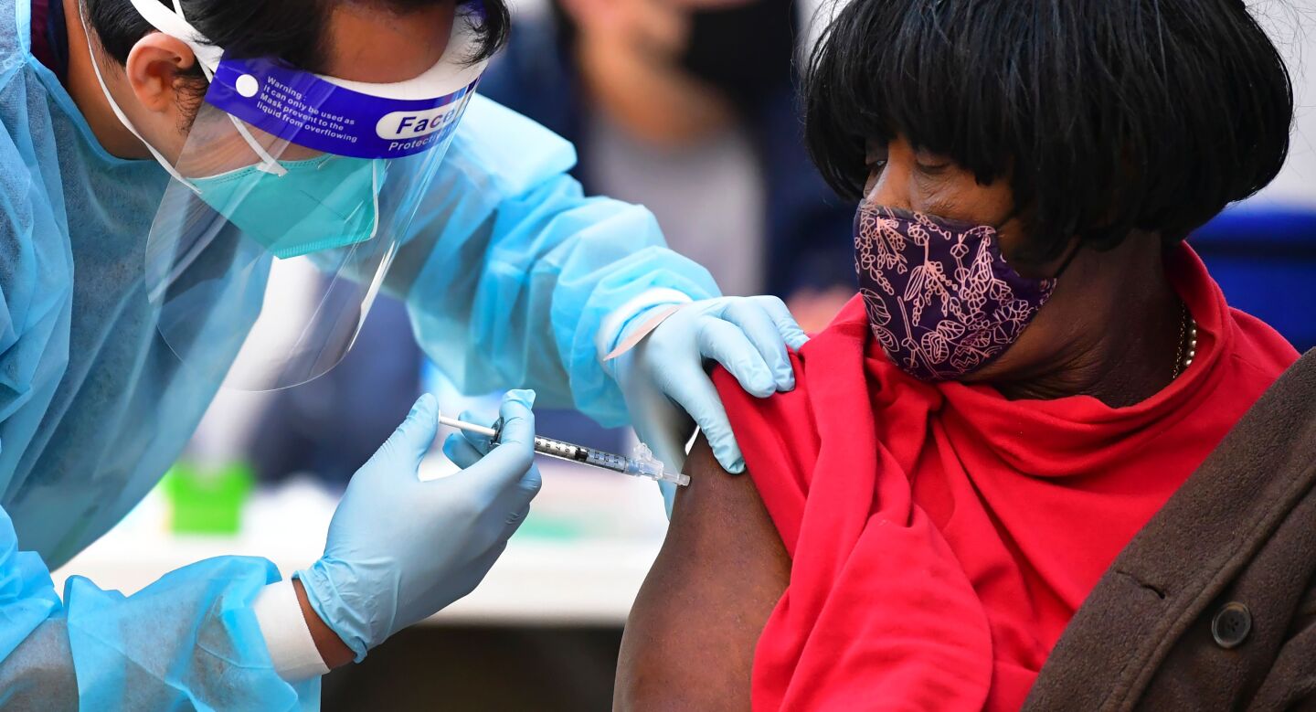 A nurse in blue personal protective gear administers the Moderna Covid-19 vaccine to a woman in a red shirt.