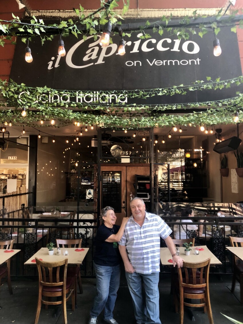 Dora Herrera and Ermanno Neiviller pose for a photo in front of the Il Capricco storefront. The two are standing and Dora has her hand placed on Ermanno's shoulder as she smiles up at him. The two stand amongst an outdoor seating area with soft patio lights and green vines strung above. 