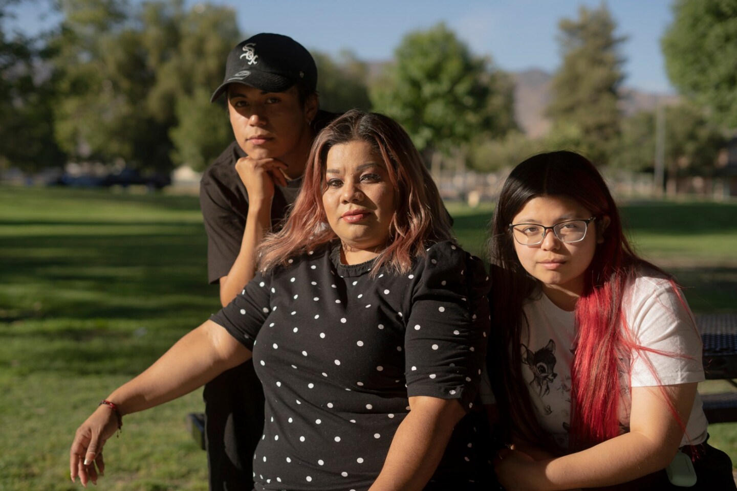 A young olive-skinned boy and girl, the boy wearing all black and a black cap, the girl in glasses wearing her pink-tipped hair long and loose, sit outside with their mother between them, who also has pink tipped hair and a black shirt with white dots.