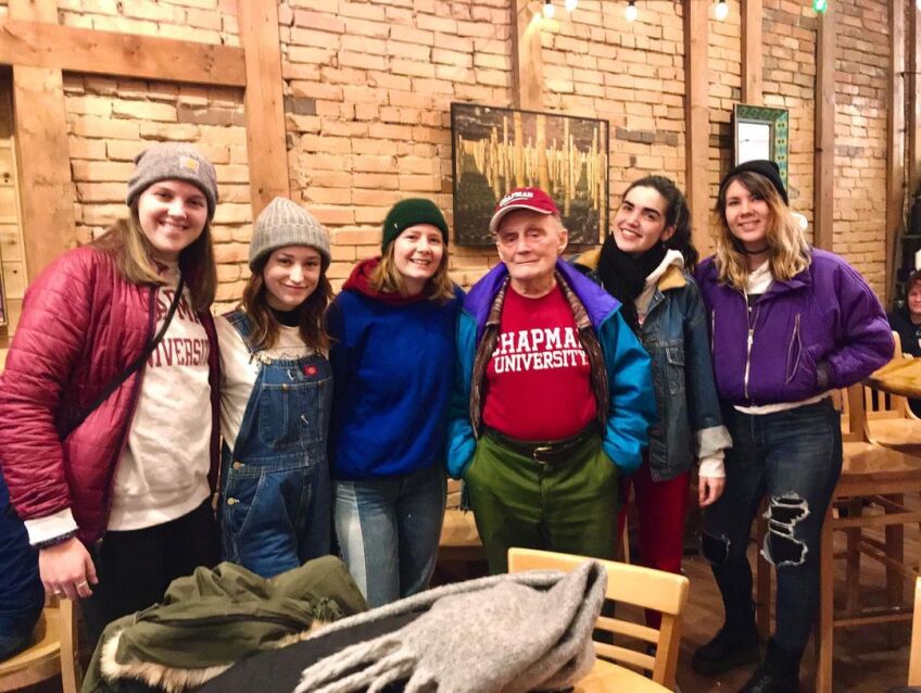 A group of college-age women pose for a photo next to an elderly man wearing a bright red sweater that reads, "Chapman University." They are in a cafe with warm lighting and an exposed brick wall behind them.