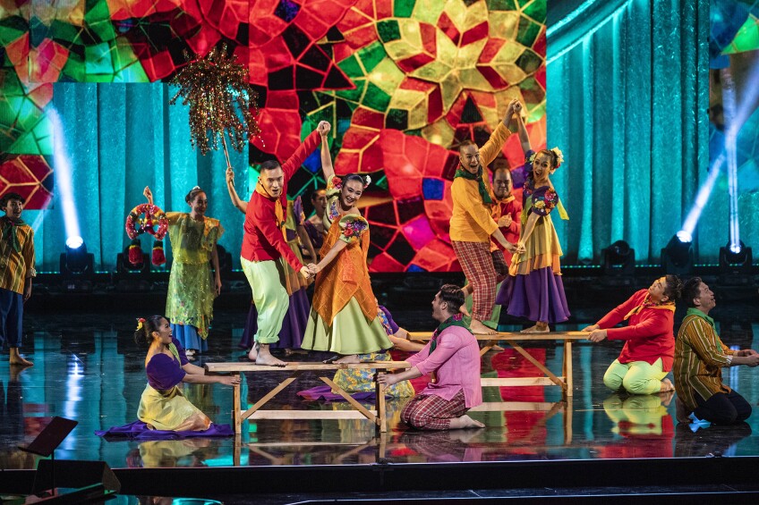 People of Southeast Asian descent perform on stage wearing brightly colored dresses and shirt-pants combinations. Two sets of couples stand on a wooden bench, facing each other while holding hands and turning to the audience with a joyous expression. People kneel on either side of the benches, holding it for stability. Behind the performance is a brightly colored mosaic of stars.