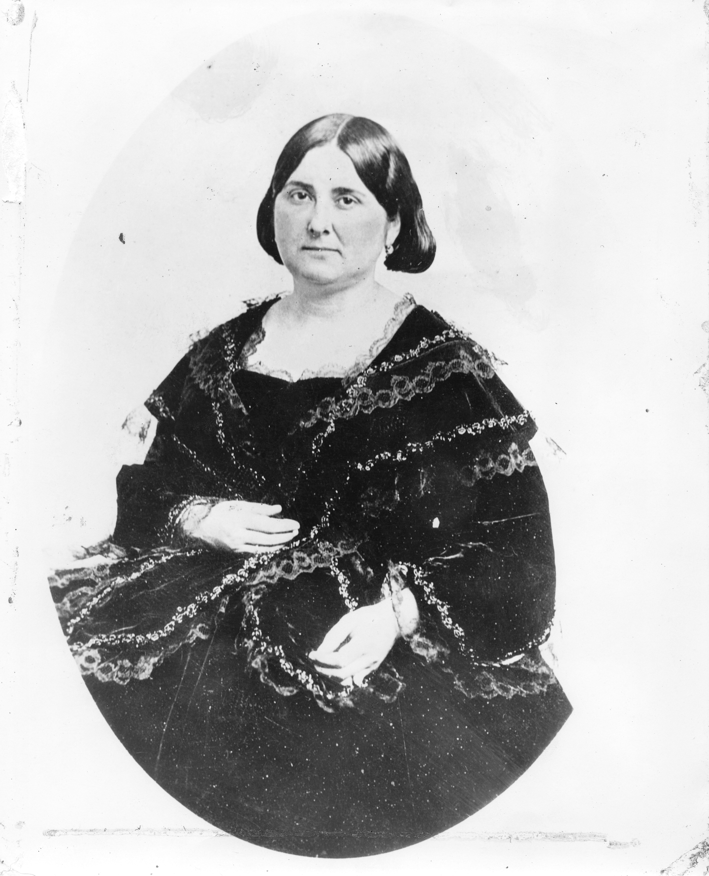 Photographic portrait of Mrs. Arcadia de Baker; previously Mrs. Abel Stearns, Arcadia Bandini, ca.1885. She can be seen from the waist up turned slightly to the left in an oval cutout. Her long dark hair is parted up the middle and pulled back to her neck. She is wearing a frilly shawl over a frilly dress with a low neckline.