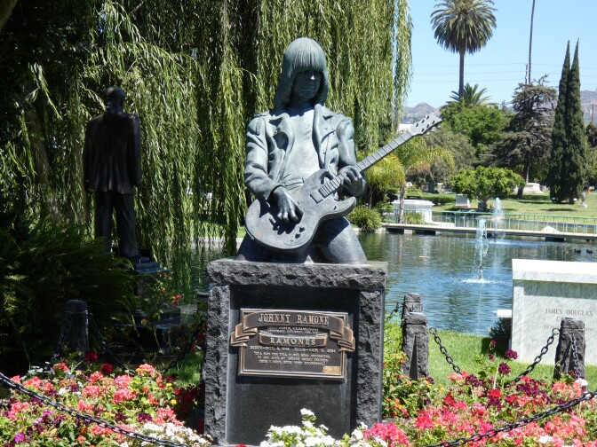 A bronze statue that has turned a gray-ish green from oxidation is a sculpture of Johnny Ramone. He has long hair and is kneeling as he plays an electric guitar. Below, a plaque reads: Johnny Ramone, Ramones. 
