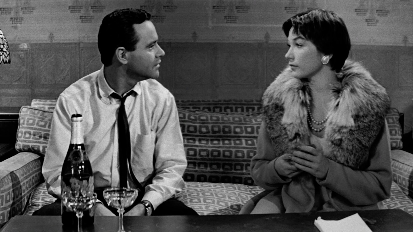 A man and woman look at each other while sitting on a couch behind wine glasses and a bottle.