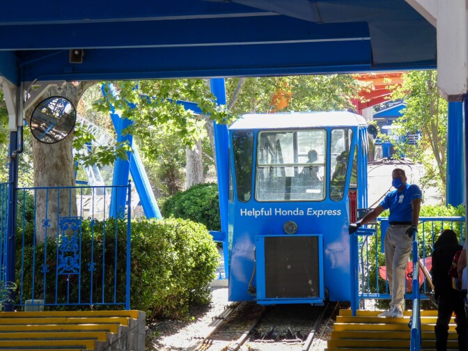 An operator in a royal blue polo shirt and beige khakis leans against the control panel as the royal blue funicular comes down the ramp, arriving at the station. The front of the funicular reads, "Helpful Honda Express." The shadow of two passengers can be seen beyond the glass of the funicular. People in line can be seen preparing for the oncoming funicular.