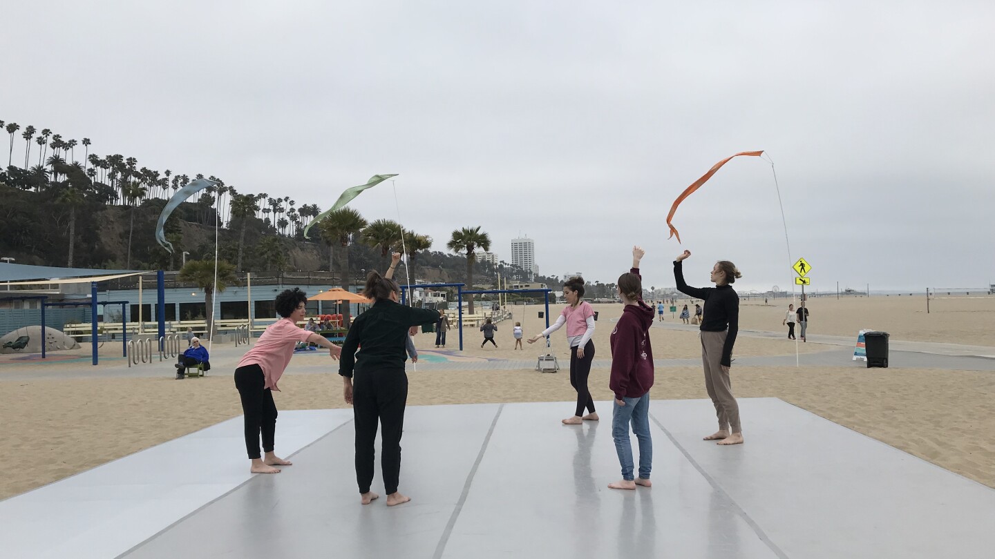 Participants stand on a platform placed on top of the sand at Santa Monica Beach. The participants are waving around different colored scarves in the air. The sky above them is overcast.