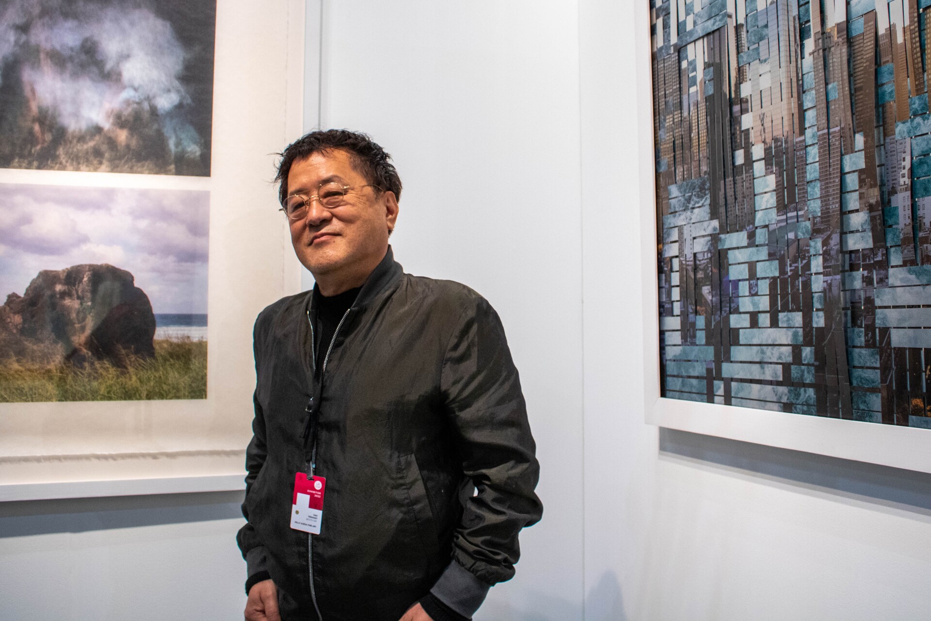Taiji Terasaki stands with his hands tucked into his pockets and looking at the camera. He's wearing a black bomber jacket. Behind him are two of his art pieces. The art piece to the left are still printed photographs of the environment. The piece on the right is woven sheet metal with environmental photos printed onto the metal.