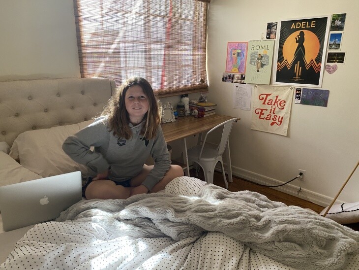 A blonde girl sits on a bed with a laptop next to her.