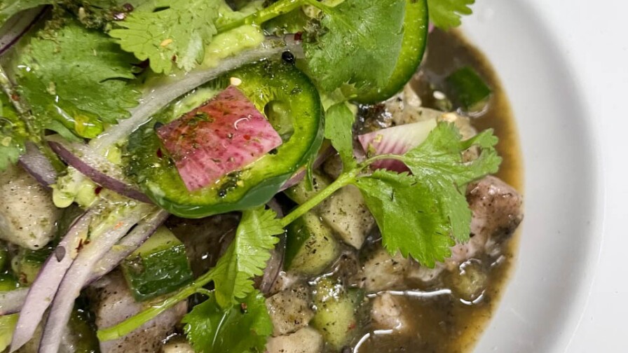 A white ceramic shallow dish holds the fish ceviche. The chopped fish is a pale gray color and sits in a dark liquid sauce. Mixed in the ceviche are sliced red onions, cucumbers, jalapeños, and cilantro.