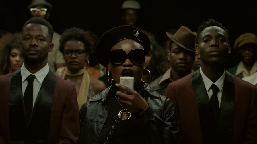 Still from Pongo's "Tamulya": A woman with a beret, leather jacket, sunglasses and a PA microphone, with men in suits on either side and others in the background