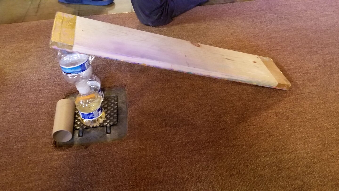A small ramp on a carpet made out of a wood slat and propped up with two small water bottles