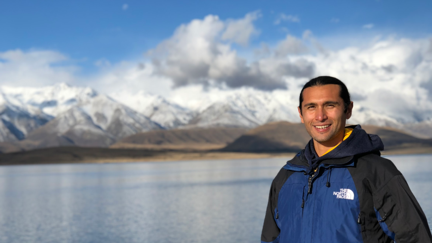 A man in a blue jacket smiles in front of a lake.