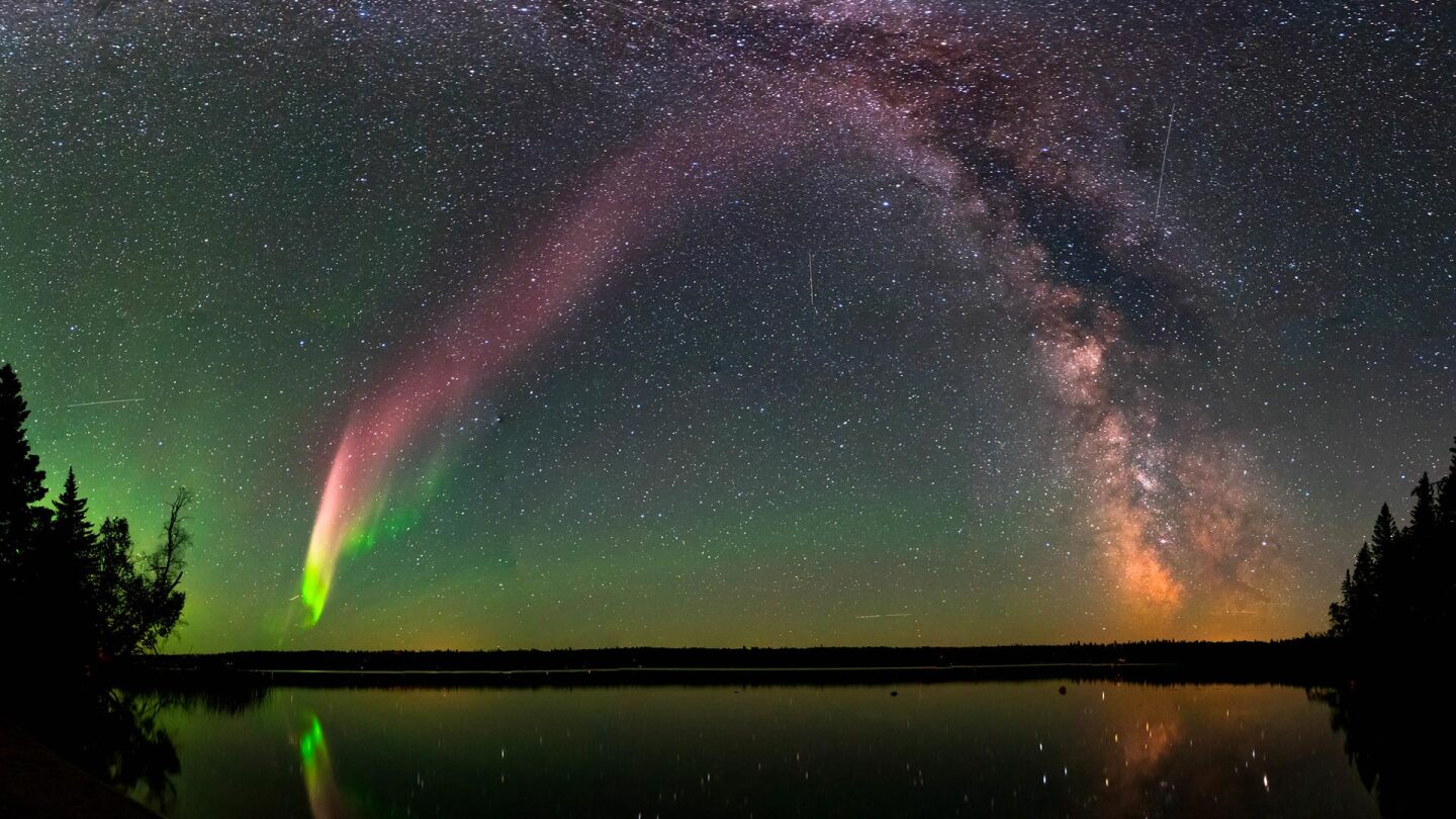 An aurora borealis and the Milky Way in the sky above a forest and lake.