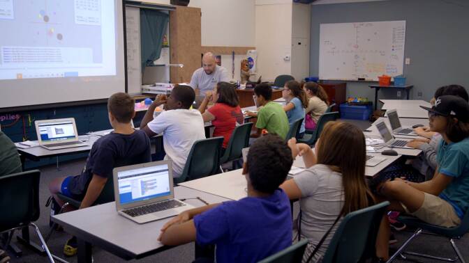 Students sit in a classroom with laptops on their desks.
