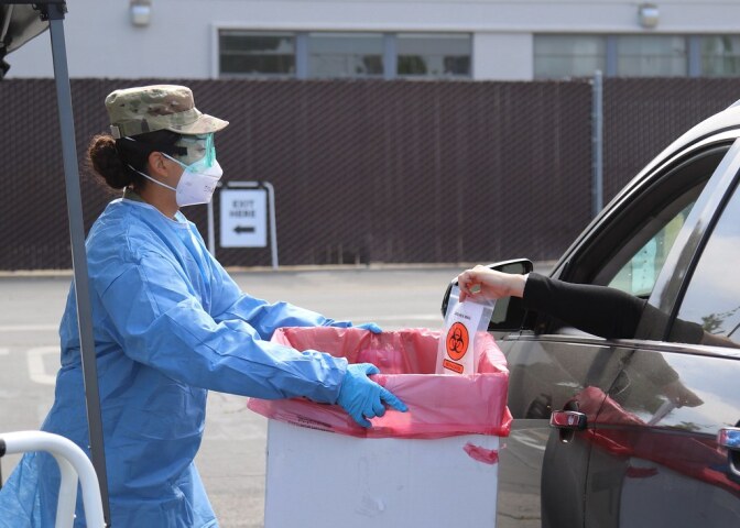 A member of the national guard in blue protective gear holds a box out to a person placing a coronavirus test in a box from a car.