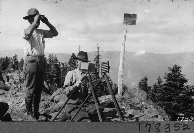 A man uses mirrors of a heliograph to flash fire messages. Meanwhile, a man standing next to him looks through binoculars.