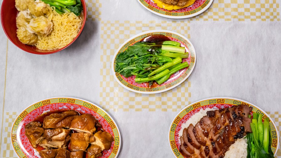 A flat lay photo of dishes from Pearl River Deli over a checkered yellow and white tablecloth. In the top left corner is a bowl of yellow noodles with leafy greens and siu mai. In the bottom left corner is a plate of roasted poultry in a dark brown sauce. In the middle is a plate of blanched leafy greens with a thick brown sauce drizzled over. In the bottom right corner is a plate of red char siu pork served over fluffy white rice and a side of leafy vegetables. At the top right corner is a plate holding a yellow bolo bao with breaded bone-in pork chop sandwiched between its buns.