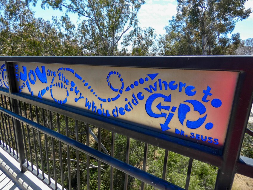 A bridge railing features decorative panels of blue plexiglass that reads, "You are the guy who'll decide where to go." Underneath that reads, "Dr. Seuss." 