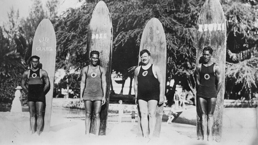 Four surfers stand in front of their boards- black and white