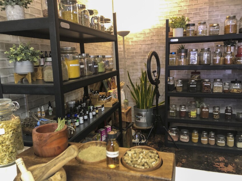 A room's walls are lined with two shelves stocked with glass jars of various sizes and shapes and filled with different powders, grains and dried leaves. A warm yellow lamp illuminates the room from the far left corner. Plants are dotted sporadically throughout the room — in the corner, on different shelves.