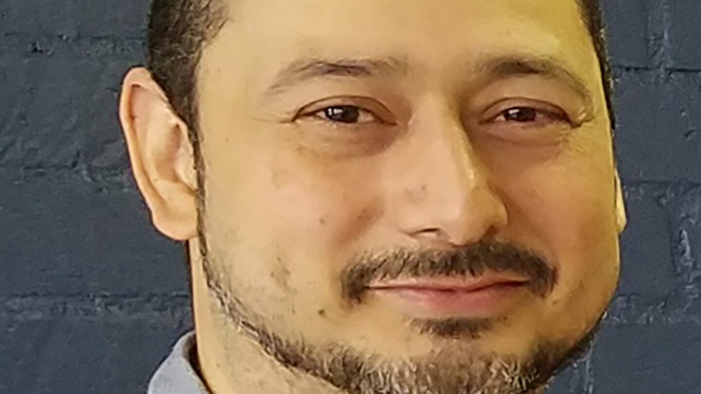 Carlos Aguilar smiles while wearing a grey button-down shirt.