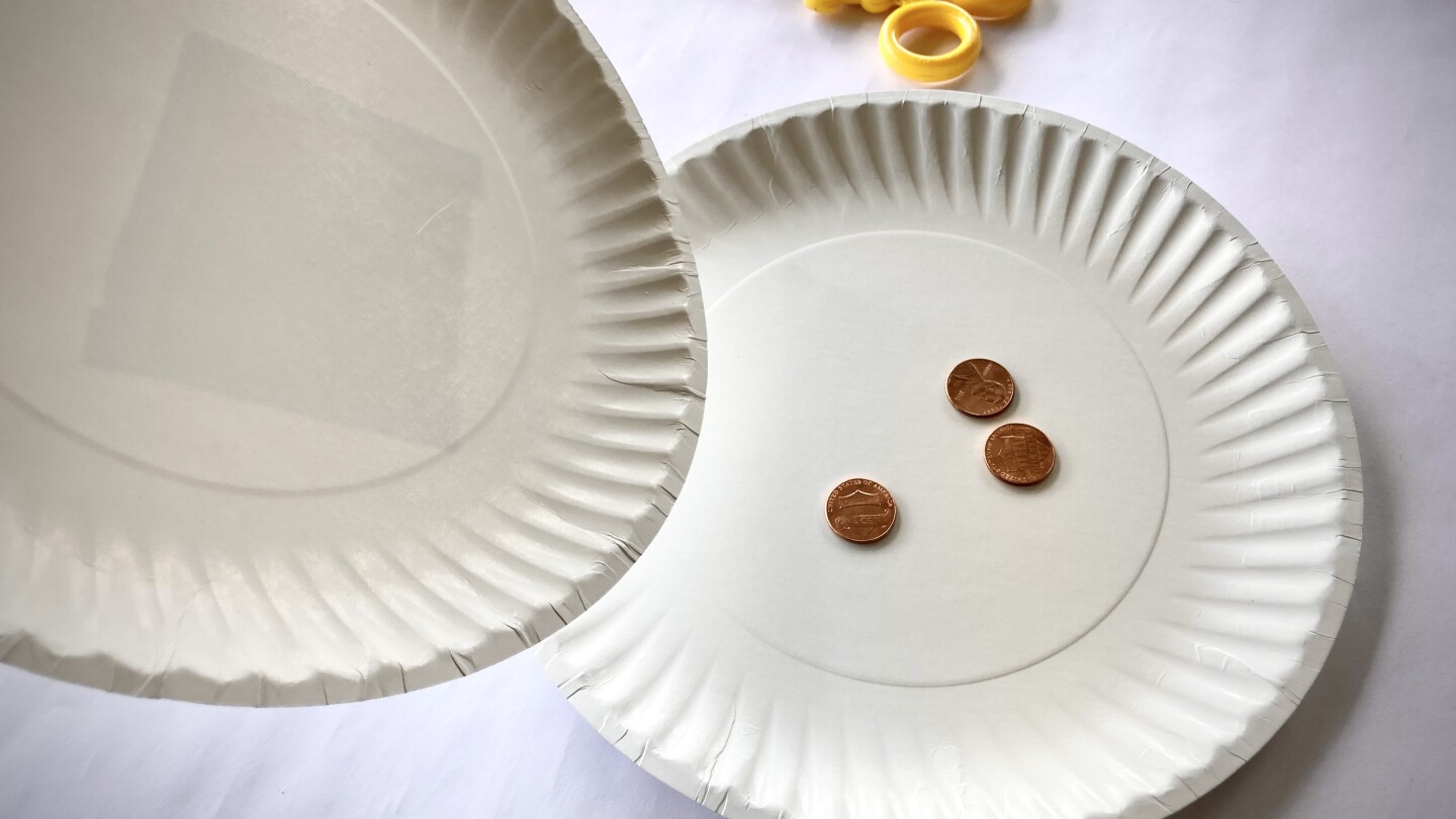One empty paper plate is tilted over another that holds three pennies on a table.