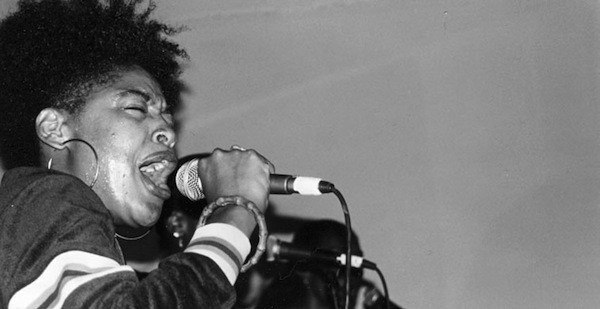 A black and white hpoto of Medusa The Gangsta Goddess, a young Black woman with her hair in an updo and wearing large hoop earrings. She is holding a microphone up to her mouth as she performs passionately into it.
