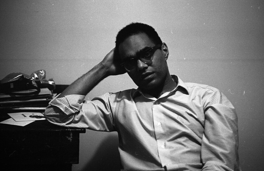 Portrait Of Robert Parris Moses in black and white. He is leaning his elbow against a table and holding his head in his hand. He is a Black man in a button down shirt and black glasses.