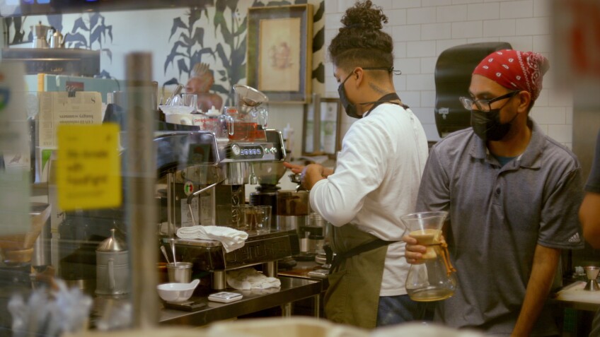 Two baristas work in a kitchen, preparing coffee. One barista has a red bandana tied around their head and holds a Chemex while the other is working at an espresso machine. Both baristas are wearing black surgical face masks.