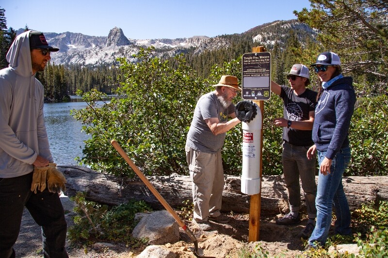 Three people gather around a trail post with a pipe attached for trash. Beyond, is a beautiful scene of a lake in front of a snow-capped mountain range. A man in the left enters the frame, holding a pair of work gloves.