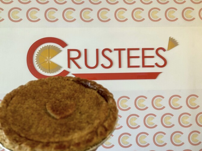 A small, single-serving pie is held up against a wall with the Crustees logo on it. The Crustees logo is red. Inside the C is an icon of a pie with one slice missing. A slice of pie takes the place of the apostrophe by the 'S.'