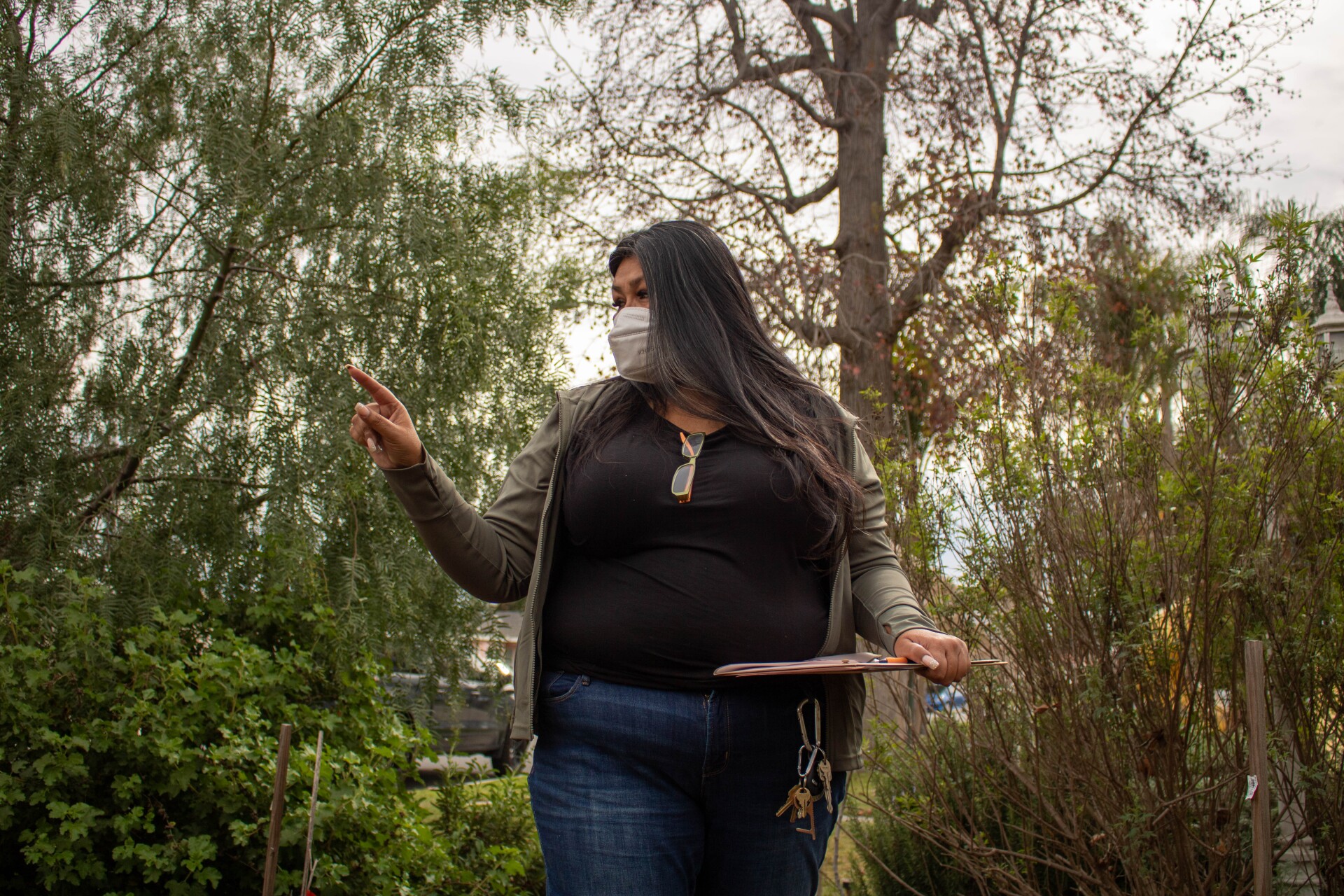 A woman in a black t-shirt and gray sweater points to her right while holding a clipboard. She is looking around inquisitively.