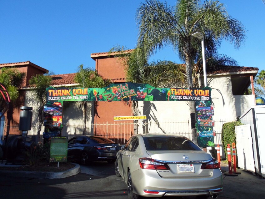 Cars drive through the entryway for a tiki-themed car wash. a sign hangs above the entrance that reads, "Thank you! Please enjoy the ride!" and "Tiki Tour Express Wash."