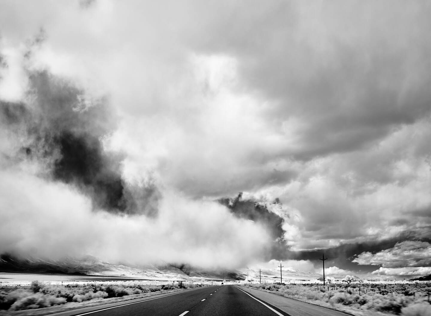 A black and white photograph of a wide desert road against a cloudy sky.
