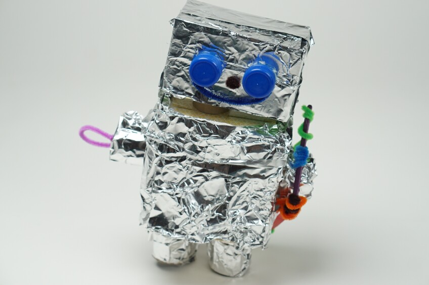 a mustachioed robot craft made out of boxes covered in aluminum foil decorated with small bottle caps for eyes and small, colorful craft pipe cleaners for arms