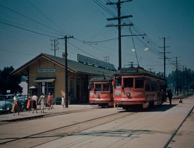 Red trolley cars are parked outside of the North Hollywood depot, a yellow paneled building with dark brown trim and a large sign across the roof that reads "Southern Pacific - Pacific Electric Station." Pedestrians approach the depot on the sidewalk.