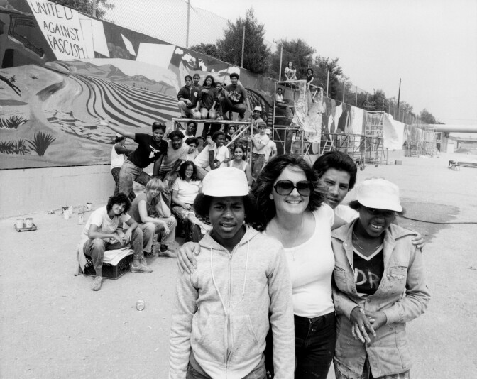 A black and white photo features a group photo with Judy Baca standing in the foreground wearing glasses and her arms wrapped around youth artists. Behind them are the other youth artists, gathered around scaffolding. They're standing in the Tujunga Wash with the "Great Wall of California" mural behind them.