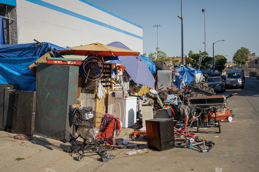 A homeless encampment consisting of umbrellas, tarps, a shopping cart, a washing machine and other debris spills out from the sidewalk and into the street, in front of parked cars.